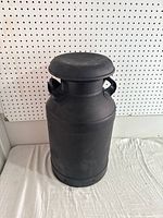 Full view of the old black metal milk can sitting on a fabric surface, against pegboard background, showing overall shape and design.