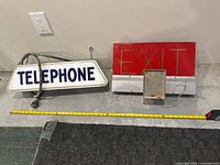 Photo showing both the TELEPHONE and EXIT electric signs side by side with measuring tape for scale