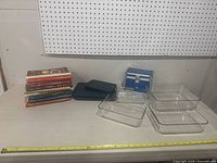 Image showing four clear glass Pyrex dishes of varying sizes, a stack of vintage cookbooks, a blue metal container, and a green plastic folder all arranged on a countertop.