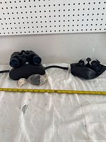 Both pairs of black binoculars with accessories and carrying cases shown side by side on a white cloth with measuring tape in view.