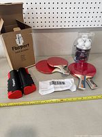 Lot contents including four red rubber paddles, container of white balls, FbSport retractable net closed and packaging box on shelf with pegboard background.