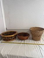 Full view of three wicker baskets arranged side by side on a cloth surface against a pegboard wall with a measuring tape in front verifying size.