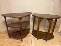 Image of two wooden demi lune tables placed side by side against a wall on a wooden floor. One table has three half-moon shelves with turned legs; the other has an octagonal top and vertical supports with a lower shelf.