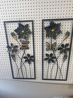 Pair of rectangular metal framed wall decorations hanging on pegboard. Each piece has black metal flowers with gold trim and butterflies, showing dimensional metalwork with some wear.