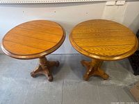 Two round oak side tables with four-legged pedestal bases shown side by side on a gray floor against a white wall, displaying wood grain and finish.
