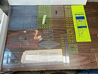 Full spread of 16 mixed quilting and sewing rulers and templates laid out on a table in various sizes and colors including translucent green, yellow, and clear plastic. Shows mix of square, rectangle, triangle, and curved shapes.