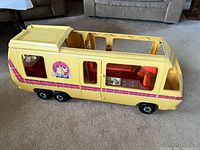 Side view of a yellow Barbie Camper/RV playset with pink striping and Barbie logo decal on the side. Windows and door openings visible. Wheels attached.