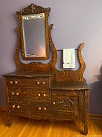 Front view of antique quarter-sawn tiger oak washstand with ornate crest, original swivel mirror, three drawers with wooden knobs, cabinet door with carved medallion, and towel bar with towel attached.