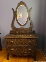 Full front view of antique tiger oak dresser with two drawers, original brass cup pulls, scalloped apron, and oval swivel mirror with decorative crown fixed on top.