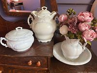 Three-piece Victorian white ironstone wash set displayed on wooden cabinet with floral decoration in background.