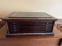 Front view of the vintage wooden flat file cabinet showing 7 black-finished shallow drawers with round brass pulls and decorative carved trim base.