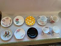Photo of assorted porcelain teacups, mug, and various saucers on a plain background; features mixed floral and polka dot patterns.