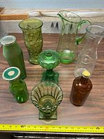 Photo shows eight assorted vintage green and amber glassware items on a wooden surface with a tape measure for size reference. Items include textured vases, pedestal pieces, pitchers, and bottles with lids.