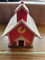 Front view of red and cream colored plastic Fisher-Price Play Family Schoolhouse with yellow clock face and red clock hands, door opening, and floral graphics.