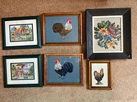 All 6 framed needlepoint and cross stitch art pieces laid on carpet showing variety of cottage and rooster designs.