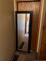 Full-length rectangular mirror with a black wooden frame angled leaning against a wooden door in a carpeted room.