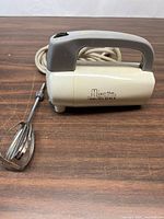 Side view of Hamilton Beach Mixette hand mixer with cord and beater attachment lying beside it on wooden surface.