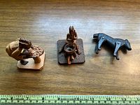 Three wood figurines placed on a wooden surface with a measuring tape showing size scale; includes two human cultural figures and one animal figure.
