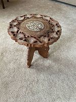 Full view of the hand-carved wooden accent table with intricate cutout design and white inlay floral pattern on top, shown standing on carpet.