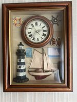 Frontal view of the nautical shadow box wall clock showing all decorative elements including mini lighthouse, sailboat, lifebuoy, ship wheel, and sailor's pulleys inside a wood frame with glass front.