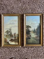 Pair of vertical oil paintings with landscape scenes, both in ornate gold frames, seen from above on carpet.