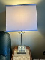 One lamp illuminated on a side table next to dark furniture, showing clear Lucite column base, square chrome platform base, and rectangular white fabric shade with minor wear.