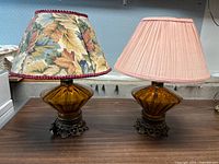Pair of vintage amber glass table lamps shown with floral and pink shades on dark metal bases, placed on wooden surface.