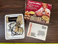 Photo of the electric food gun device placed on a tray, with power cord, next to the instruction manual and original box.
