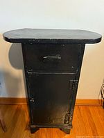Front view of vintage black wooden cabinet showing drawer and cabinet door with black handle and latch, placed on hardwood floor.