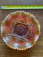 Top view showing the vivid marigold iridescent surface with Wishbone & Spatula pattern including floral and basketweave details. The dish edge is visibly ruffled, and a measuring tape above the dish shows just under 9 inches in diameter.