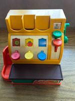 Front view of vintage Fisher Price cash register showing buttons and coin slots on yellow front panel with visible wear and missing blue button sticker.