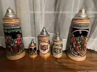Front view of the set of 5 ceramic German beer steins showing detailed relief scenes and ornate pewter lids