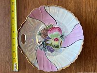 Front side of vintage decorative plate showing hand-painted fruit centerpiece with apples, grapes, pineapple, accent panels in pink and iridescent white with gold trim, plus pierced handle.