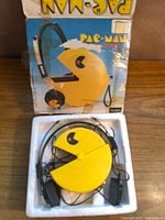 Photo of the vintage Pac-Man AM-FM radio in original white foam tray with attached headphones, displayed on wooden surface with original box in background.