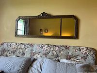 The entire vintage style rectangular wall mirror mounted on a yellow wall above a floral couch, showing bronze/antique gold ornate composite frame with decorative crest at top center.