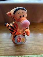 Front view of plush Tigger toy upright on a wooden surface with attached tag visible, highlighting facial features and soft fabric material.