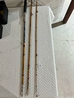 Three sections of an antique split bamboo fishing rod laid out on a white textured surface. Shows detailed rod sections, metal ferrules, line guides, and the black handle grip.