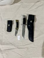 Two Buck knives pictured side-by-side on beige cloth. Folding knife with black handle and brass hardware on the left. Fixed blade knife with leather sheath on the right.