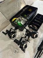 Image showing 3 Shimano spinning reels positioned in front of an open tackle box. The tackle box is filled with various fishing accessories including lures, hooks, lines, and tools.