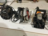 Top view showing three power tools: a circular saw, biscuit joiner, and orbital sander arranged on a white surface.