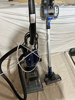 Front view of Hoover SteamVac upright vacuum and Hoover cordless stick vacuum side by side showing power brush and hose attachment on the SteamVac and slim design of cordless model