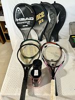 Four tennis racquets on display with their protective covers behind. Two unopened tennis ball sleeves in front.