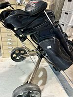 Black golf bag loaded with golf clubs, set on a black and silver golf push cart with three wheels, showing side profile.
