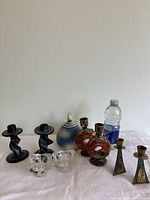 Full view of the collection showing variety of candlesticks and candleholders made of stone, decorated glass, metal, crystal, and clay arranged on white cloth with a water bottle for scale.