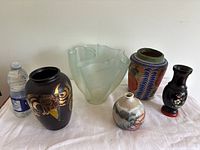 Full view of six vases including frosted glass, painted ceramic, wood and floral decorated vases next to a water bottle for scale