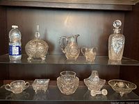A shelf displaying an assortment of crystal items including a large carafe with stopper, a large jug, a small jug, a round candy dish, small square dishes, and a vase, all with intricate cut designs.