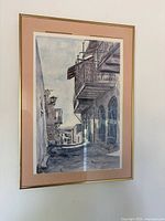 Photo of custom framed art print depicting Tsfat street scene with balconies, blue shutters, and stone buildings. Frame is gold with pink matting, with print misaligned.