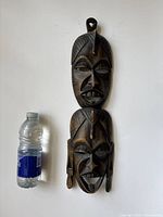 Full view of the carved double face African mask next to a bottle for size reference.