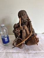 Front angle showing detailed carved face, bow, stick, bowl, and accessories including beads and metal bracelets. Water bottle for scale.