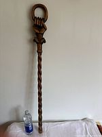 Full length view of the carved wooden staff displayed next to a water bottle for size reference. Shows twisted rope design along shaft and hand-clutch ring detail at top.